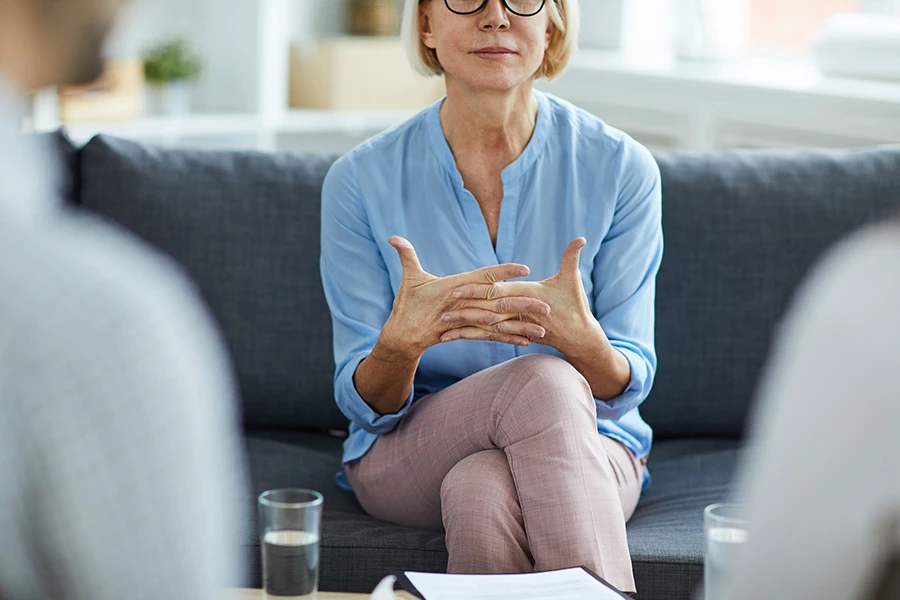 Eine Frau mit blauem Oberteil sitzt auf einem Sofa und erklärt etwas